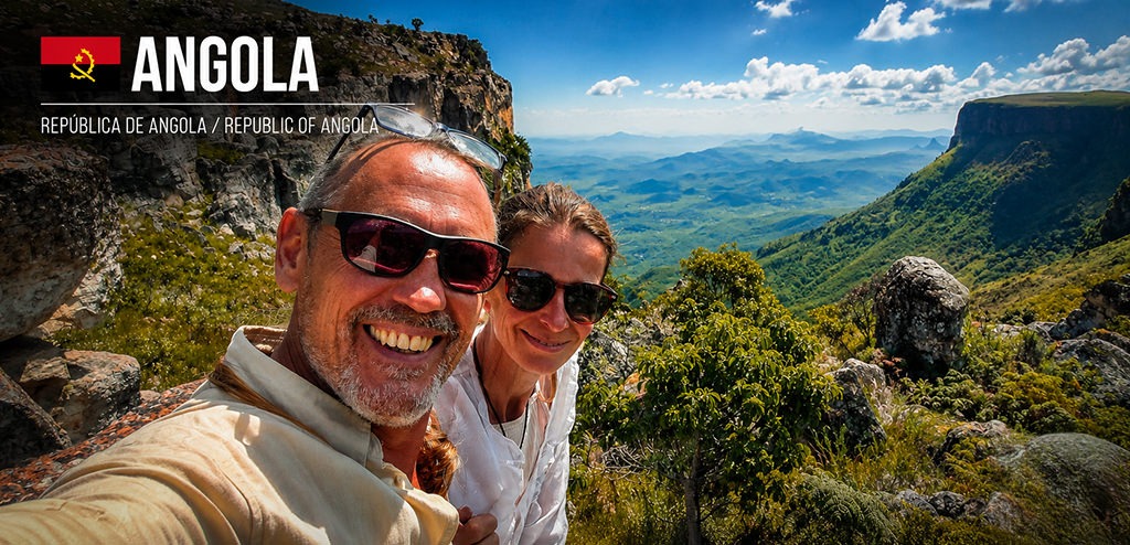 Selfie of a Fenny and me standing on a rocky mountain viewpoint in Angola, overlooking a vast valley with dramatic cliffs, rugged highlands and distant mountain ranges under a blue sky with scattered clouds.