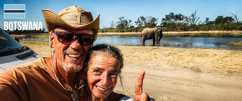 Two overlanders smiling for a selfie beside their campervan while an elephant stands at a waterhole in Botswana under a clear blue sky.