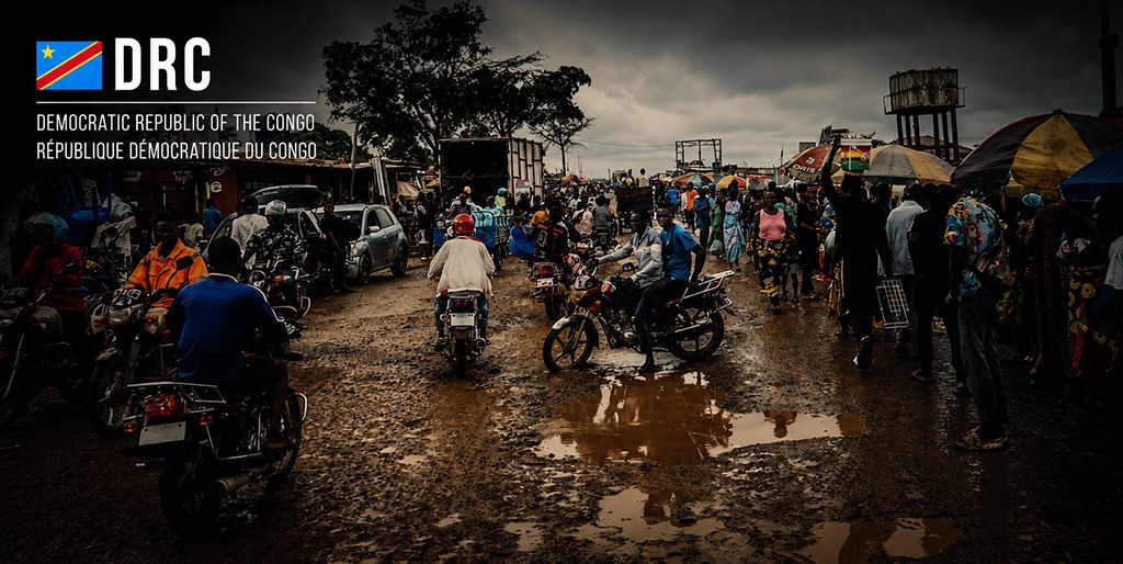 Rain-soaked street scene in the Democratic Republic of the Congo with muddy roads, dozens of motorbikes, crowded market stalls, pedestrians carrying umbrellas, and dark storm clouds gathering overhead. Country title and flag appear in the upper left corner.