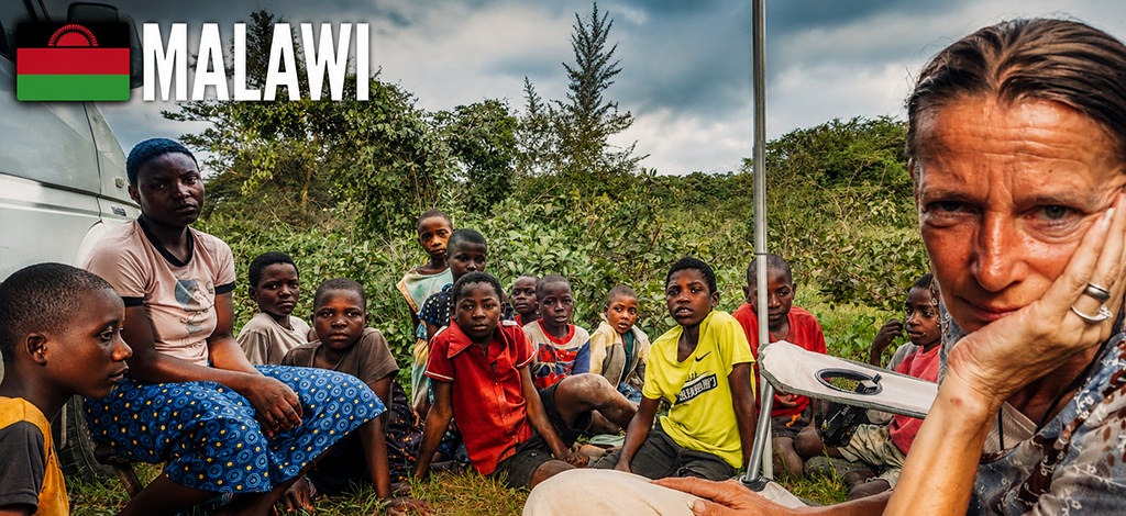 enny sitting beside Rossi in rural Malawi while a group of local children gathers around, watching curiously during a stop in the countryside.
