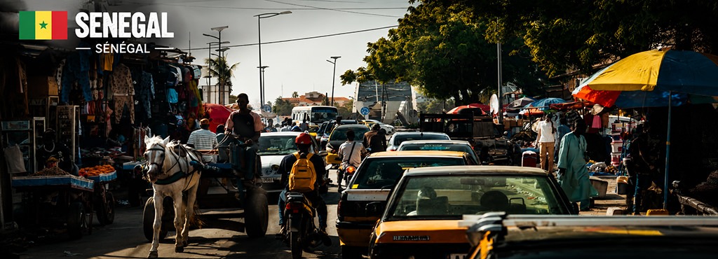 Busy street scene in Saint-Louis, Senegal, with cars, motorcycles, horse carts, street vendors and pedestrians sharing the road in a vibrant urban atmosphere.