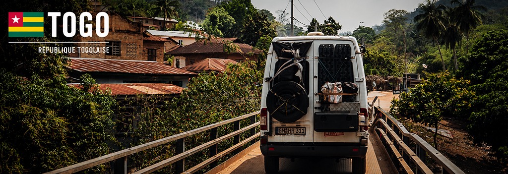 A white overland Mercedes Sprinter crosses a narrow bridge into a lush tropical village in Togo. Surrounded by green hills, palm trees and rustic houses, the scene captures the peaceful atmosphere and mountainous landscapes of one of West Africa’s smallest countries.