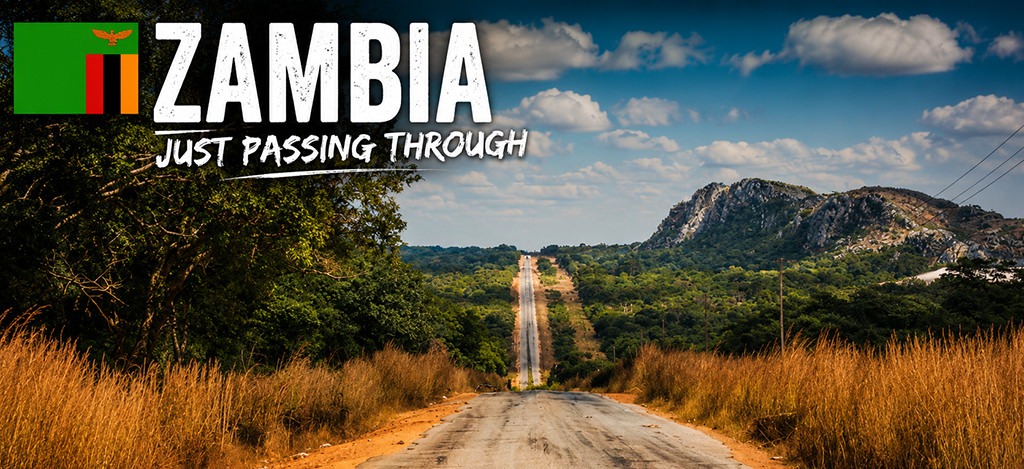 A long straight road stretches through dry grassland and woodland in Zambia, leading toward rocky hills under a partly cloudy sky.