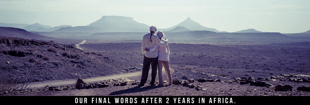 Fenny and Totti stand arm in arm overlooking a vast desert landscape with distant mountains and a winding gravel road in Namibia.