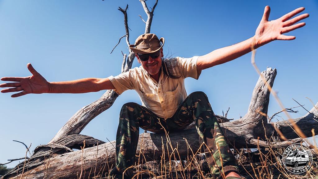 Totti sitting on a fallen tree in the African bush, welcoming the viewer with outstretched arms while sharing practical DHL Express shipping tips for overlanders.
