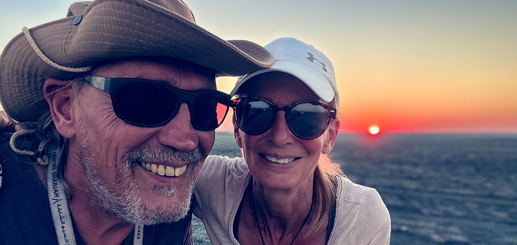 Fenny & Totti enjoying a sunset together during their full-time overlanding journey, smiling at the camera with the sun setting on the horizon.