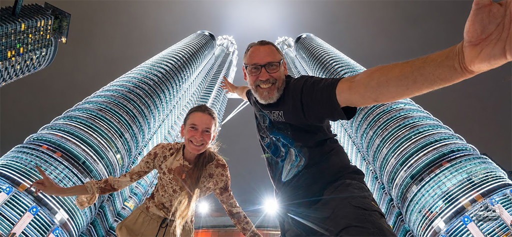 Totti and Fenny standing beneath the Petronas Twin Towers in Kuala Lumpur, photographed from a dramatic low-angle perspective.